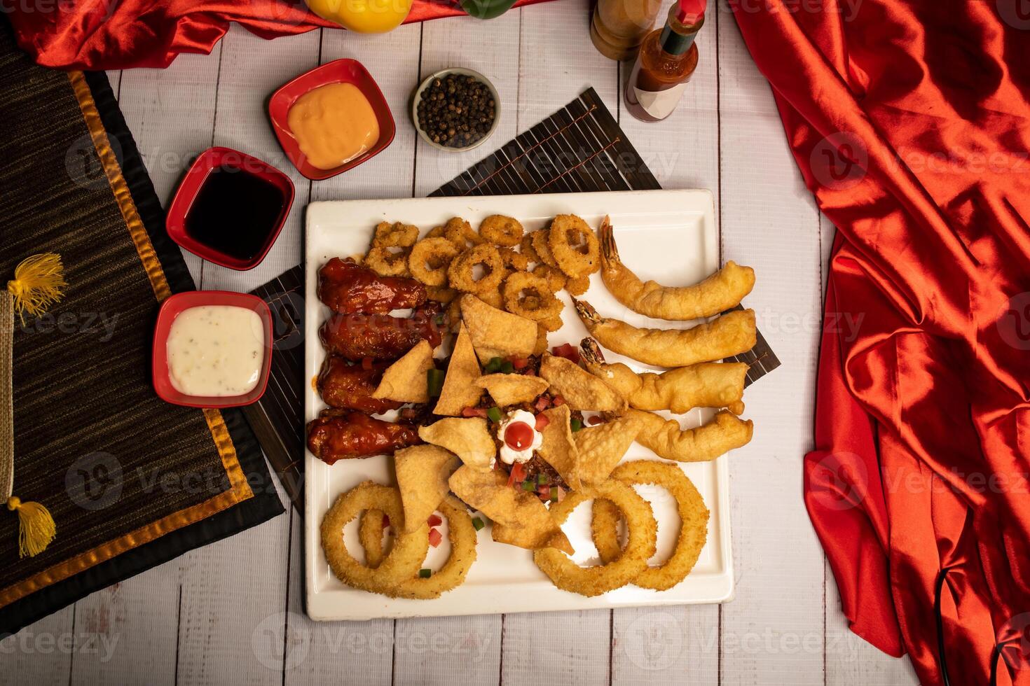 Assorted fried snacks platter with fried onion rings, shrimps, crackers, bq chicken wings, soy mayo dip sauce and black pepper served in dish isolated on napkin top view on wooden table italian food photo