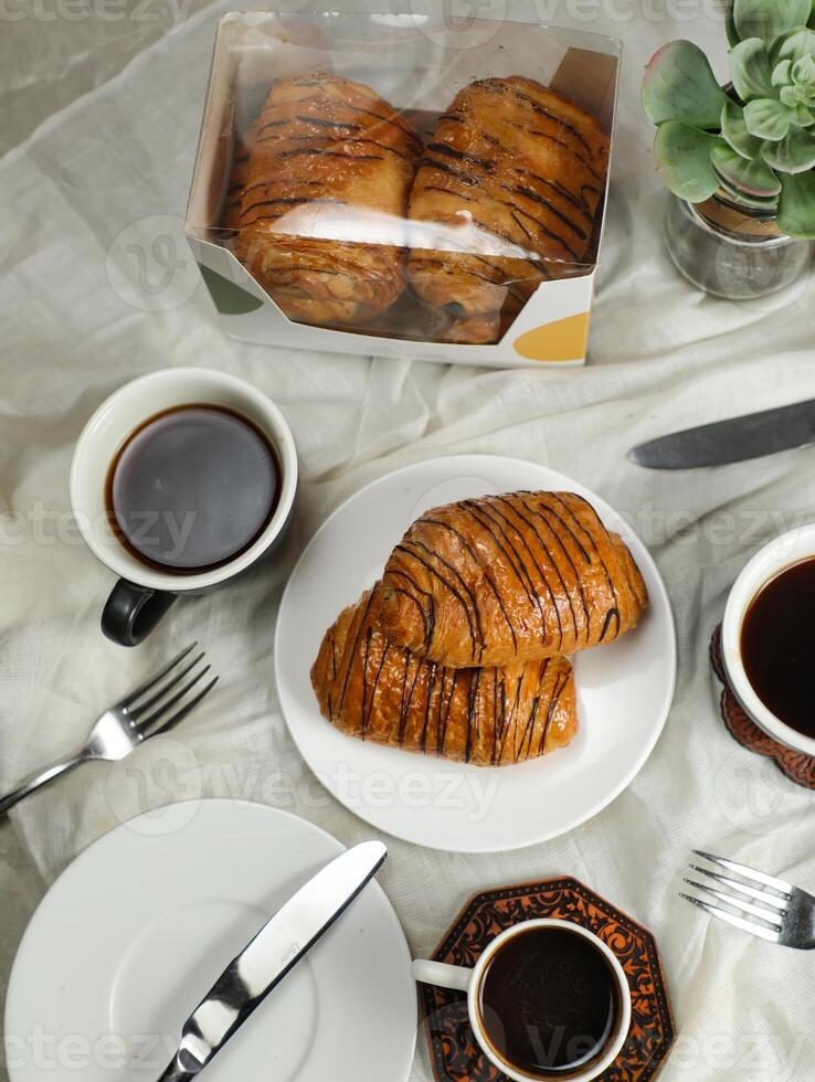 Chocolate Croissant served on plate with cup of black coffee with knife and fork isolated on napkin side view of french breakfast baked food item on grey background photo