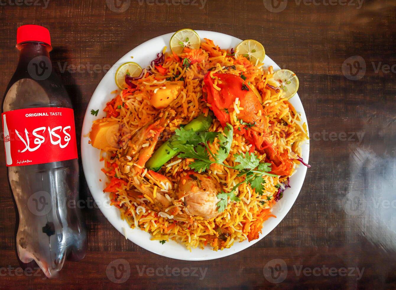 Spicy chicken biryani with potato half litter coke bottle 500 ml, onion and lemon slice served in plate isolated wooden background top view indian spices and pakistani food photo