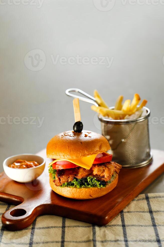 chipotle chicken burger include cheese slice, tomato, onion and lettuce leaf served on wooden board with dip and french fries bucket isolated on grey background side view of appetizer fast food photo