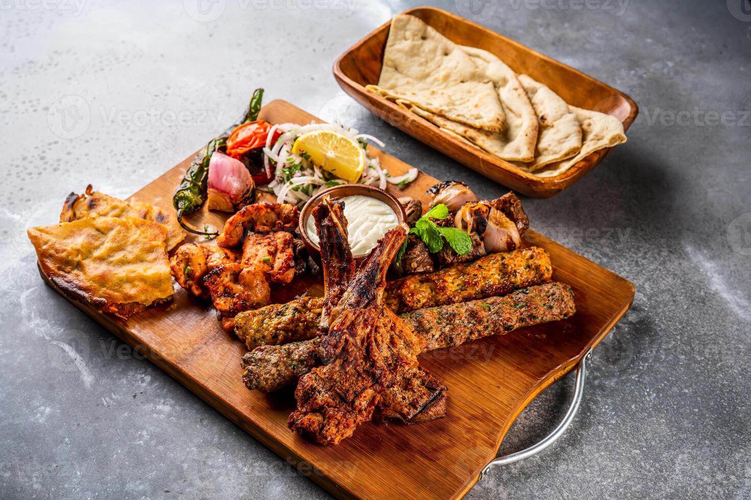 Mix grill with shish tawook, mutton chop, champ, chicken kabab, beef kebab, tikka boti, reshmi and malai pita bread, nan, tandoori roti, raita, dip, salad and lemon served in wooden board top view photo