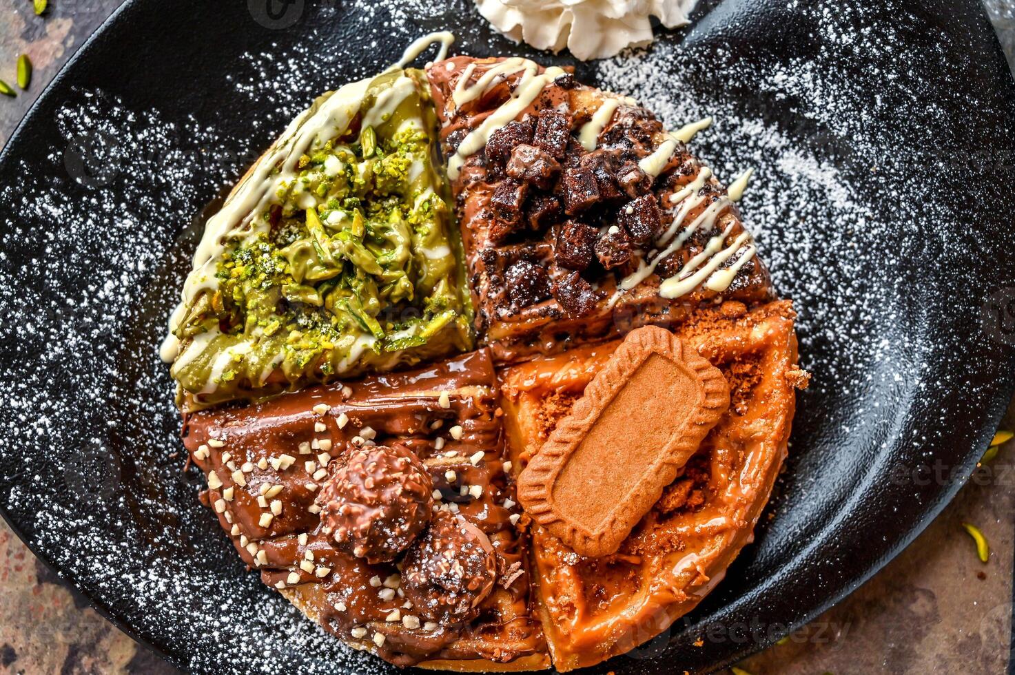 Assorted waffle with Ferrero Rocher, Nutella, Oreo, Lotus, Brownie, Pistachio, Chocolate, Whipped cream, knife and fork served in dish isolated on dark background closeup top view cafe dessert food photo