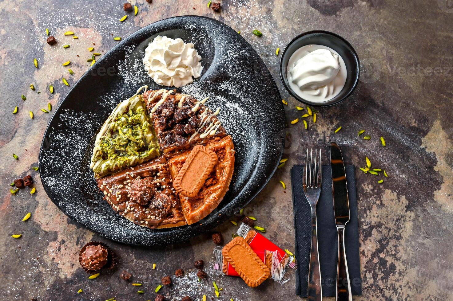 Assorted waffle with Ferrero Rocher, Nutella, Oreo, Lotus, Brownie, Pistachio, Chocolate, Whipped cream, knife and fork served in dish isolated on dark background top view cafe dessert food photo