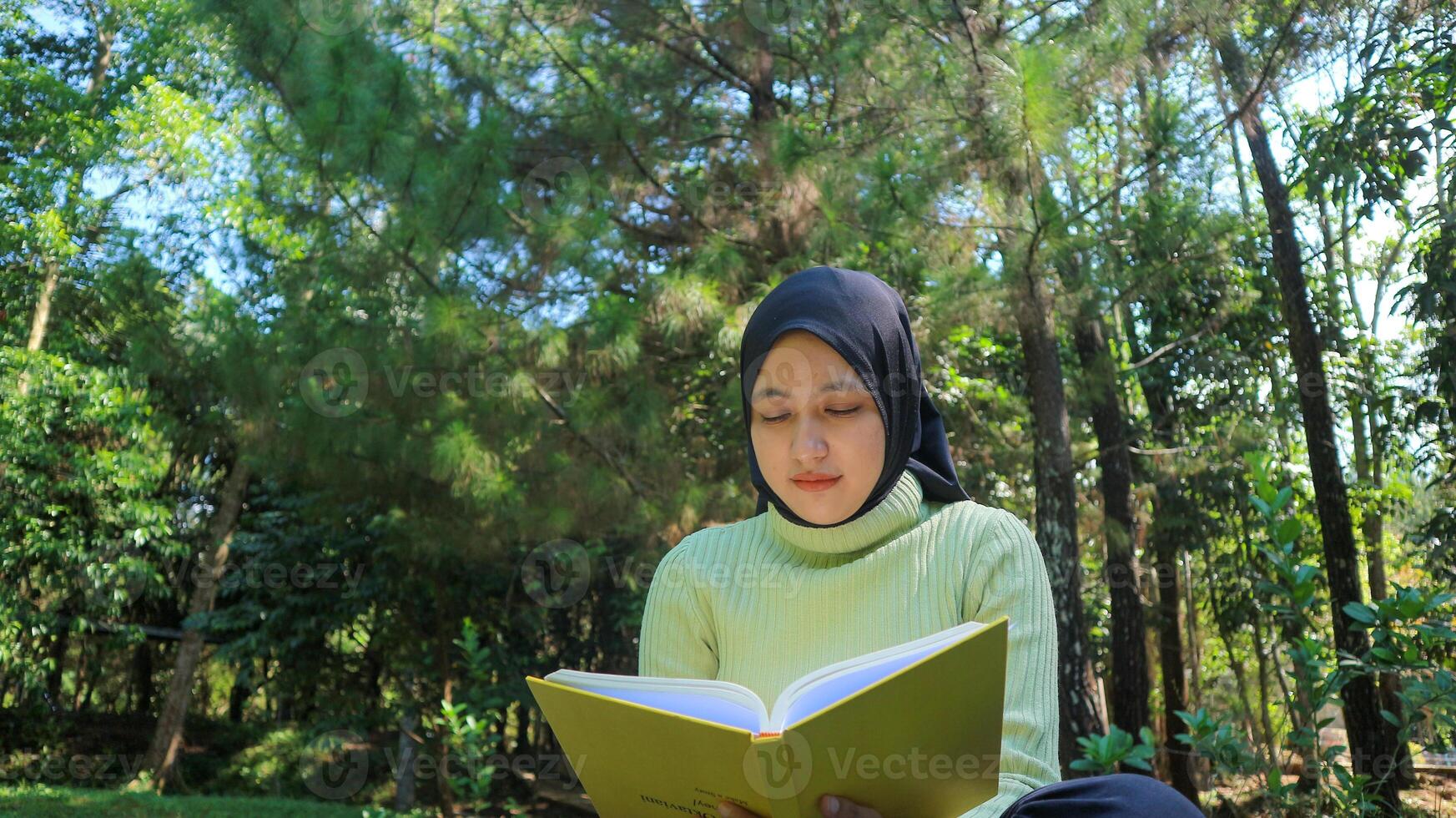 Relaxed muslim woman enjoying weekend at park, sitting on grass and reading book, empty space photo