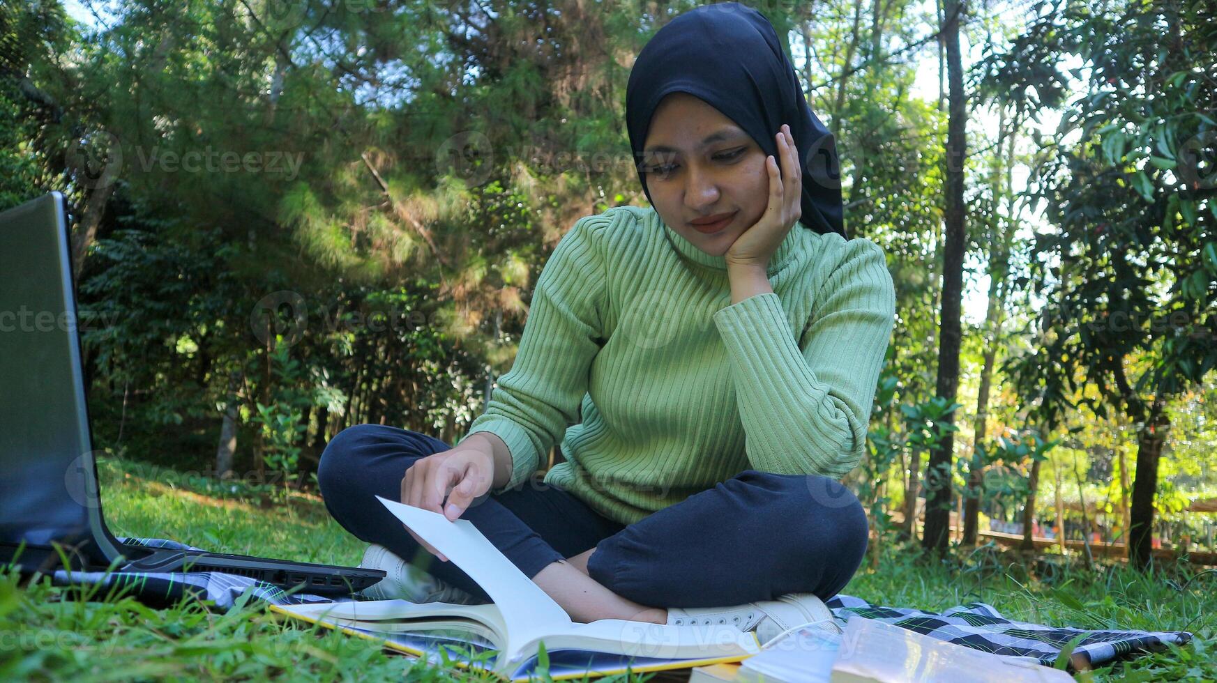 Relaxed muslim woman enjoying weekend at park, sitting on grass and reading book, empty space photo