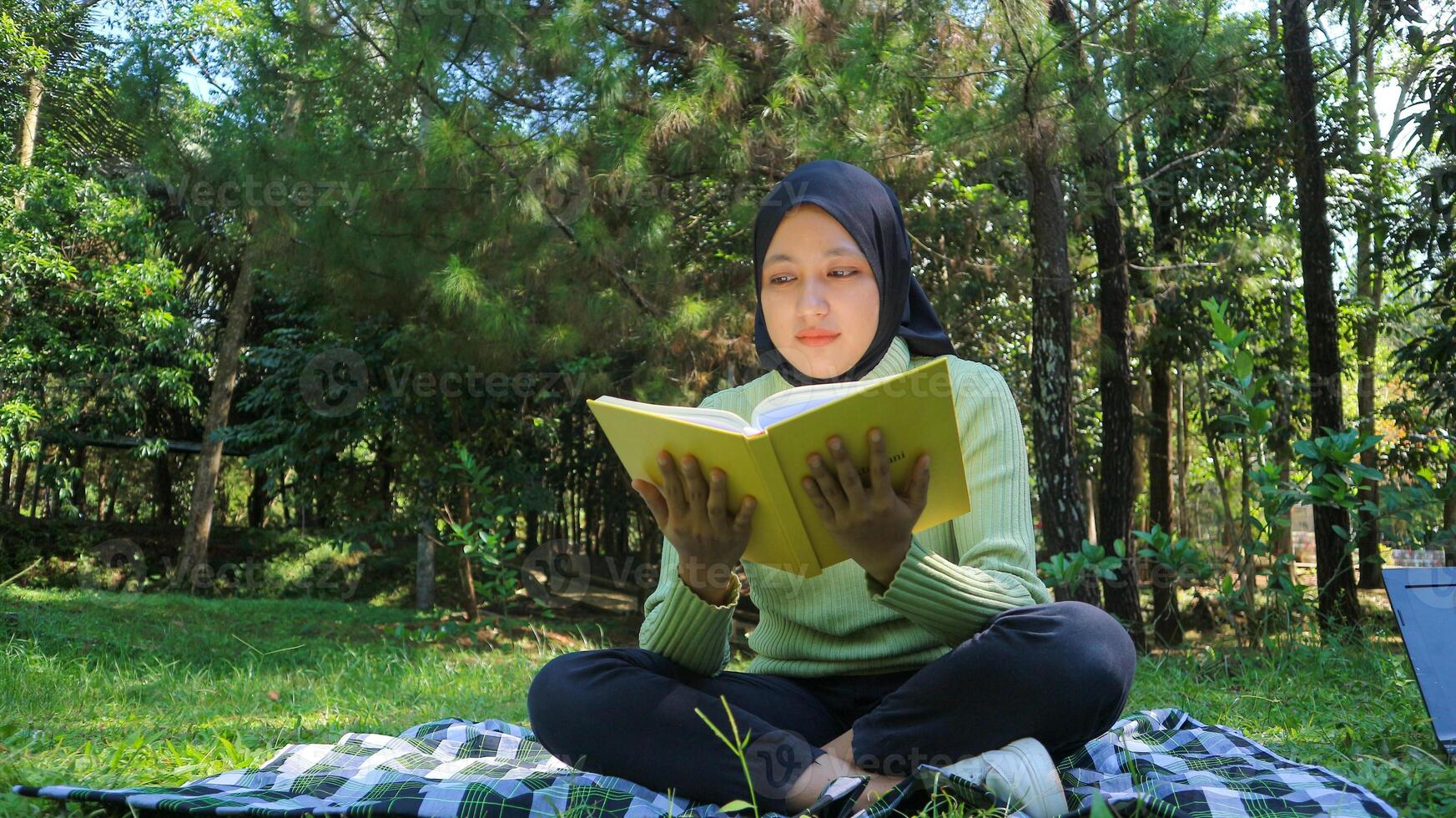 Relaxed muslim woman enjoying weekend at park, sitting on grass and reading book, empty space photo