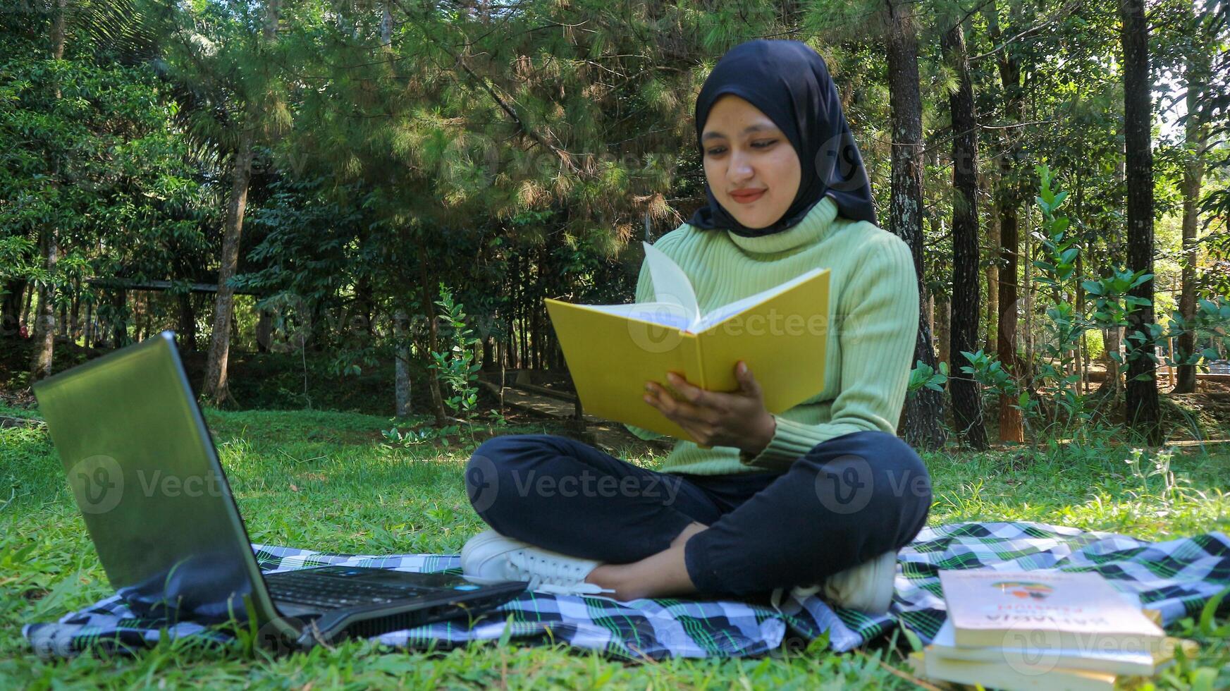 Relaxed muslim woman enjoying weekend at park, sitting on grass and reading book, empty space photo