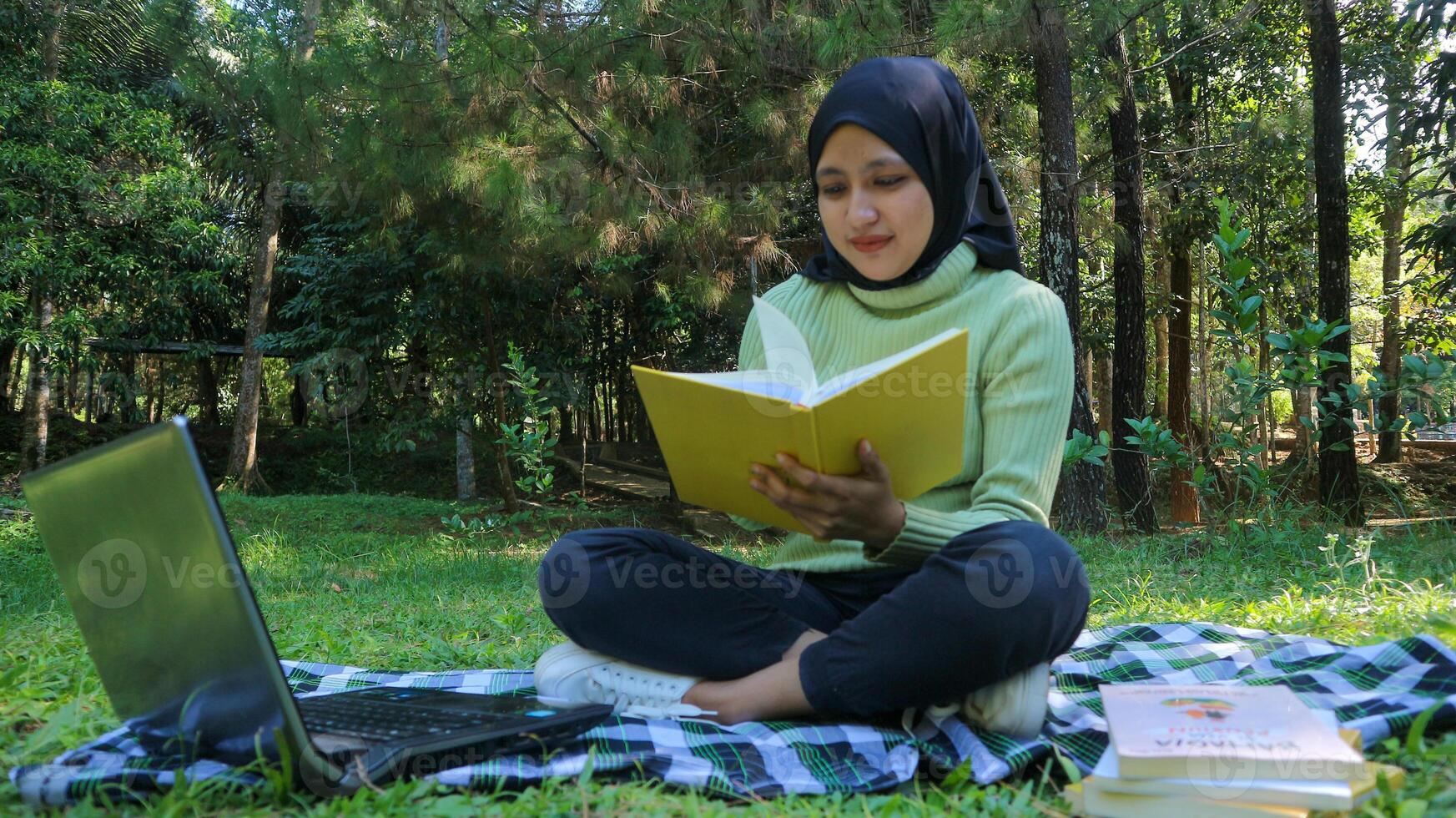 Relaxed muslim woman enjoying weekend at park, sitting on grass and reading book, empty space photo