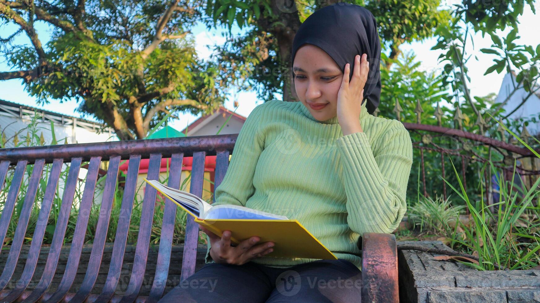 Relaxed muslim woman enjoying weekend at park, sitting on bench and reading book, empty space photo