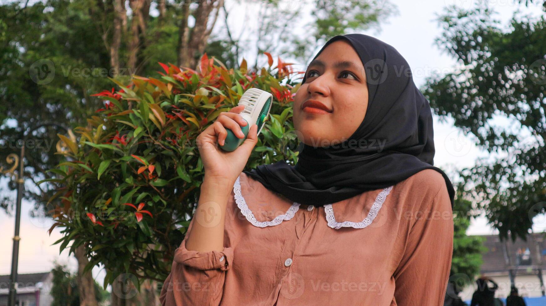 Portrait smiling of Asian muslim woman holding a portable electric fan near her face outdoors photo