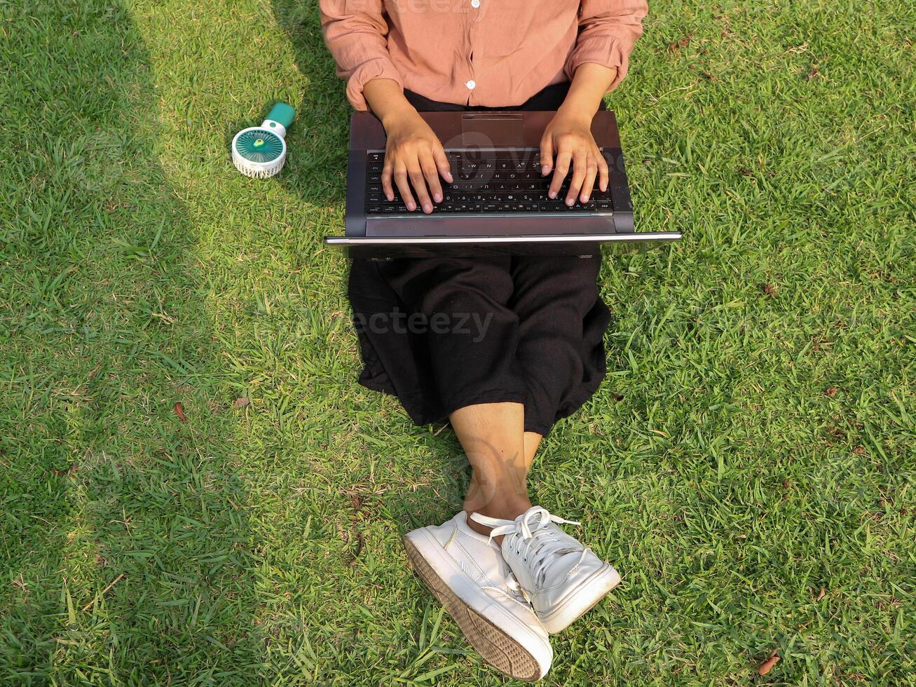 Top view of woman sitting in park on the green grass with laptop, hands on keyboard. photo