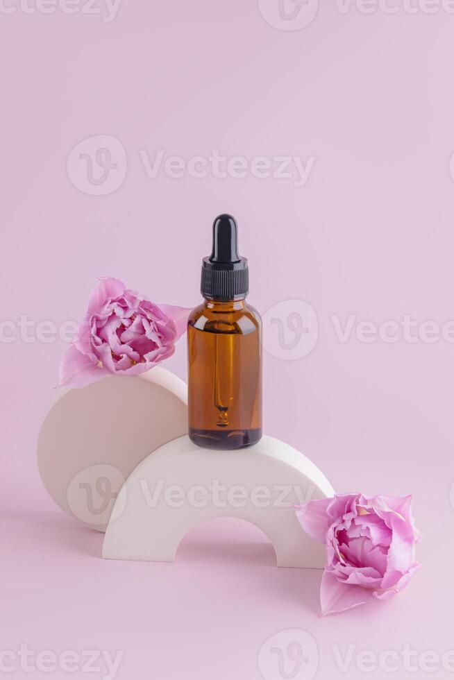 Cosmetics of nature in a glass cosmetic bottle with a pipette among concrete geometric shapes and tulip buds. Natural Cosmetics. Front vertical view. photo