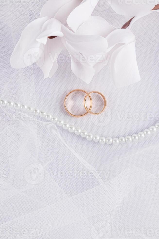 Stylish wedding vertical background of a snow-white bride's veil with two gold rings, delicate white flowers, pearl beads. postcard. invitation. photo