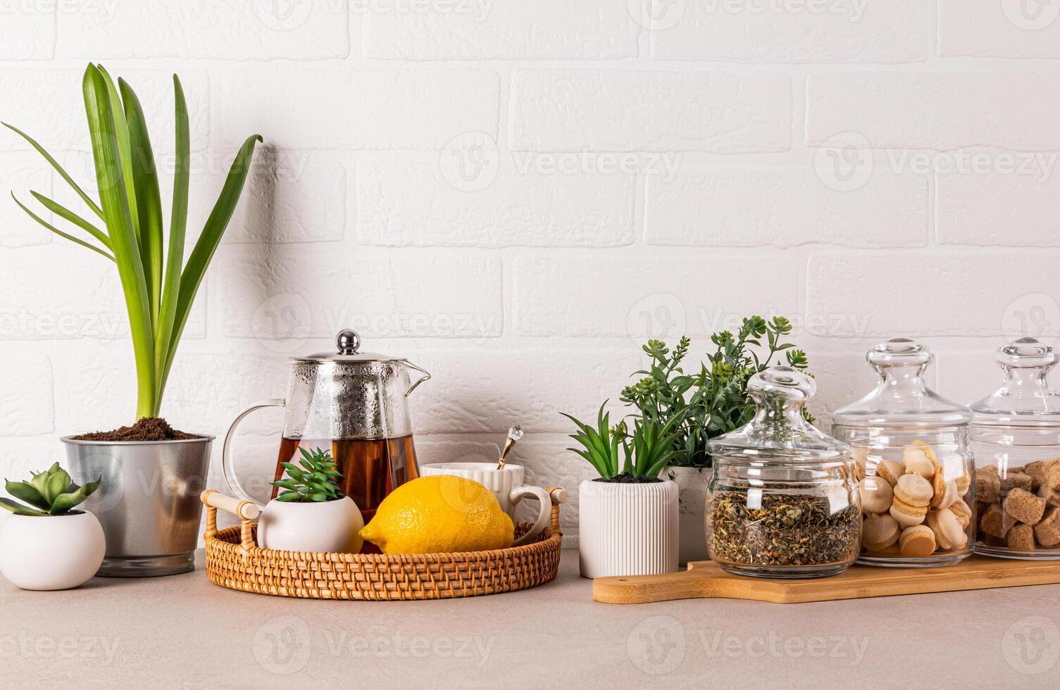 Stylish kitchen background with glass tea pot, lemon, cup, tea jars, sugar, meringue pastry. Green plants in pots. Front view. White brick wall. photo