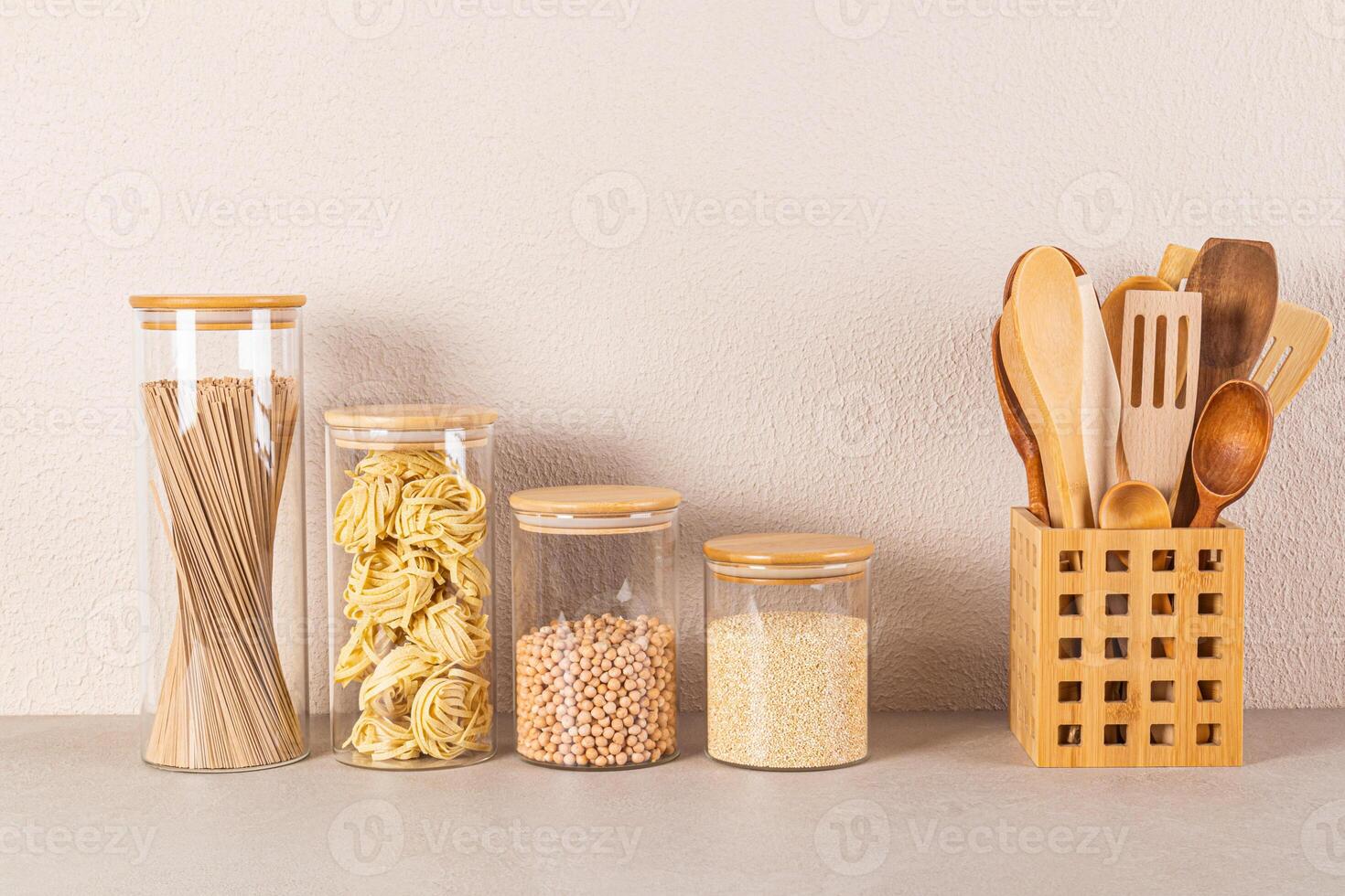 A set of glass jars with a bamboo lid for storing bulk food stand in a row next to a stand with wooden kitchen tools. Front view. minimalism. photo
