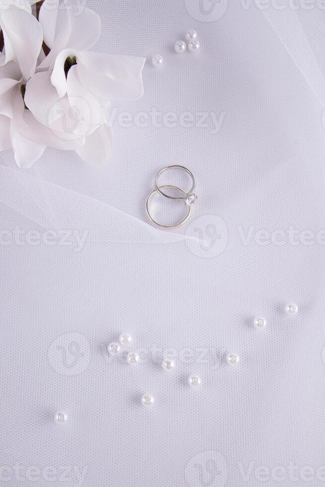 Two white gold and diamond rings lie on a veil with a scattering of pearls and flowers. Vertical wedding background. A postcard, an invitation. photo