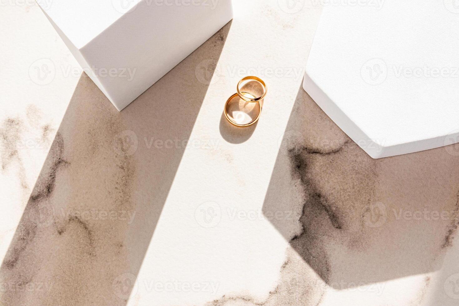 Two classic gold wedding rings lie on a marble beige background between two white geometric podiums of figures and sun rays. Top view. photo