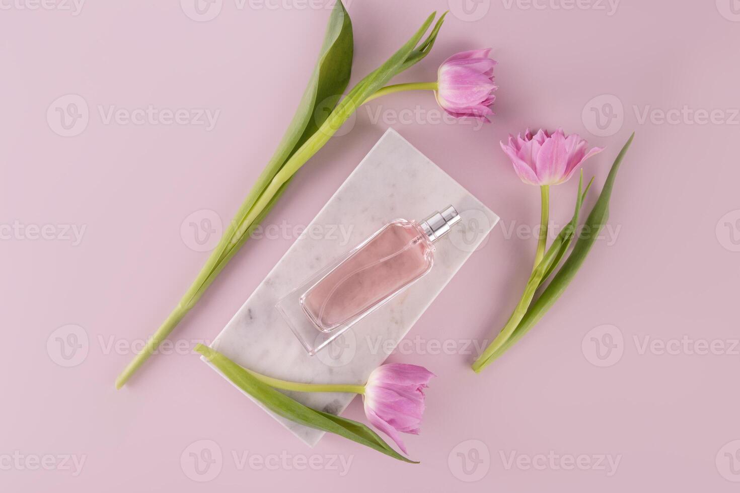 A chic bottle of women's perfume lies on a marble white podium among pink delicate tulips. Top view. Pink background. Beauty and fragrance concept. photo