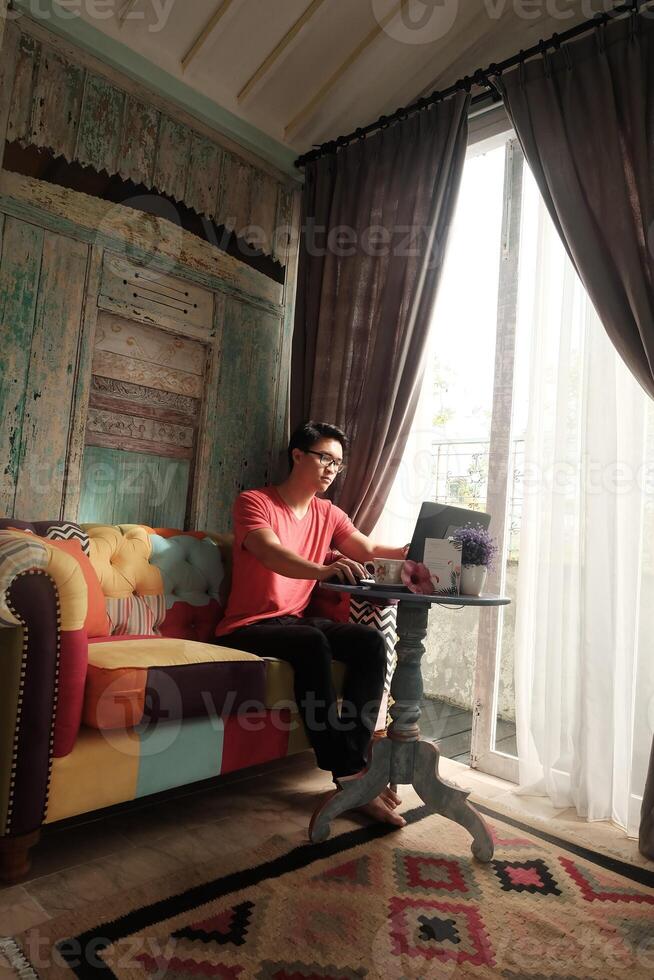 trabajo desde hogar, interior productividad, asiático hombre en sofá, trabajando en computadora portátil, con villa rayo de sol foto