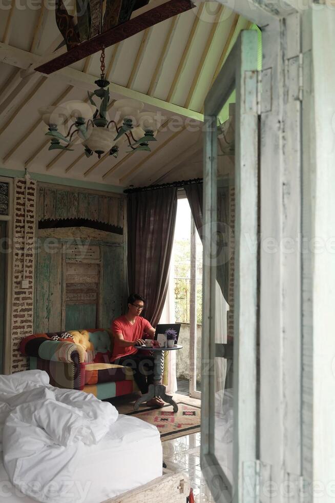 Asian Man Working on Laptop in Sunlit Villa, Sofa Comfort, Productivity photo