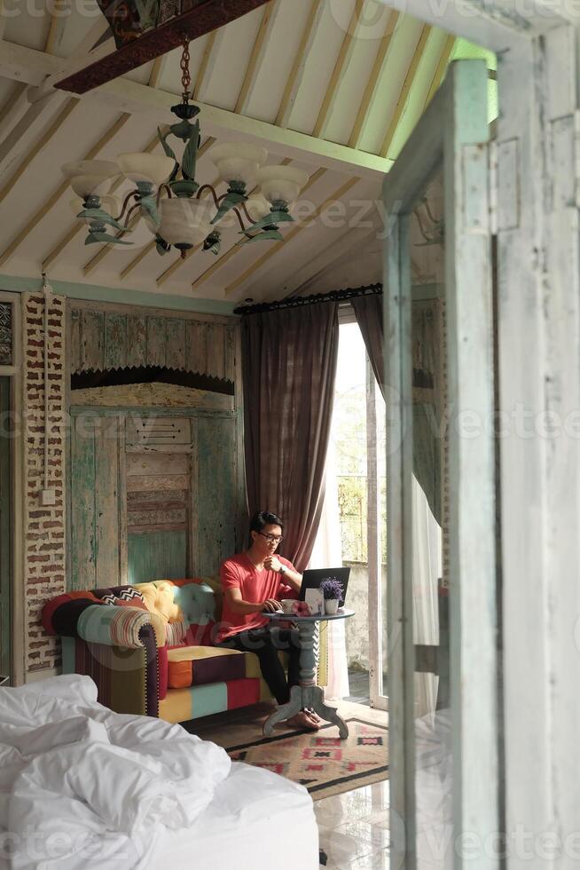 trabajo desde hogar, interior productividad, asiático hombre en sofá, trabajando en computadora portátil, con villa rayo de sol foto