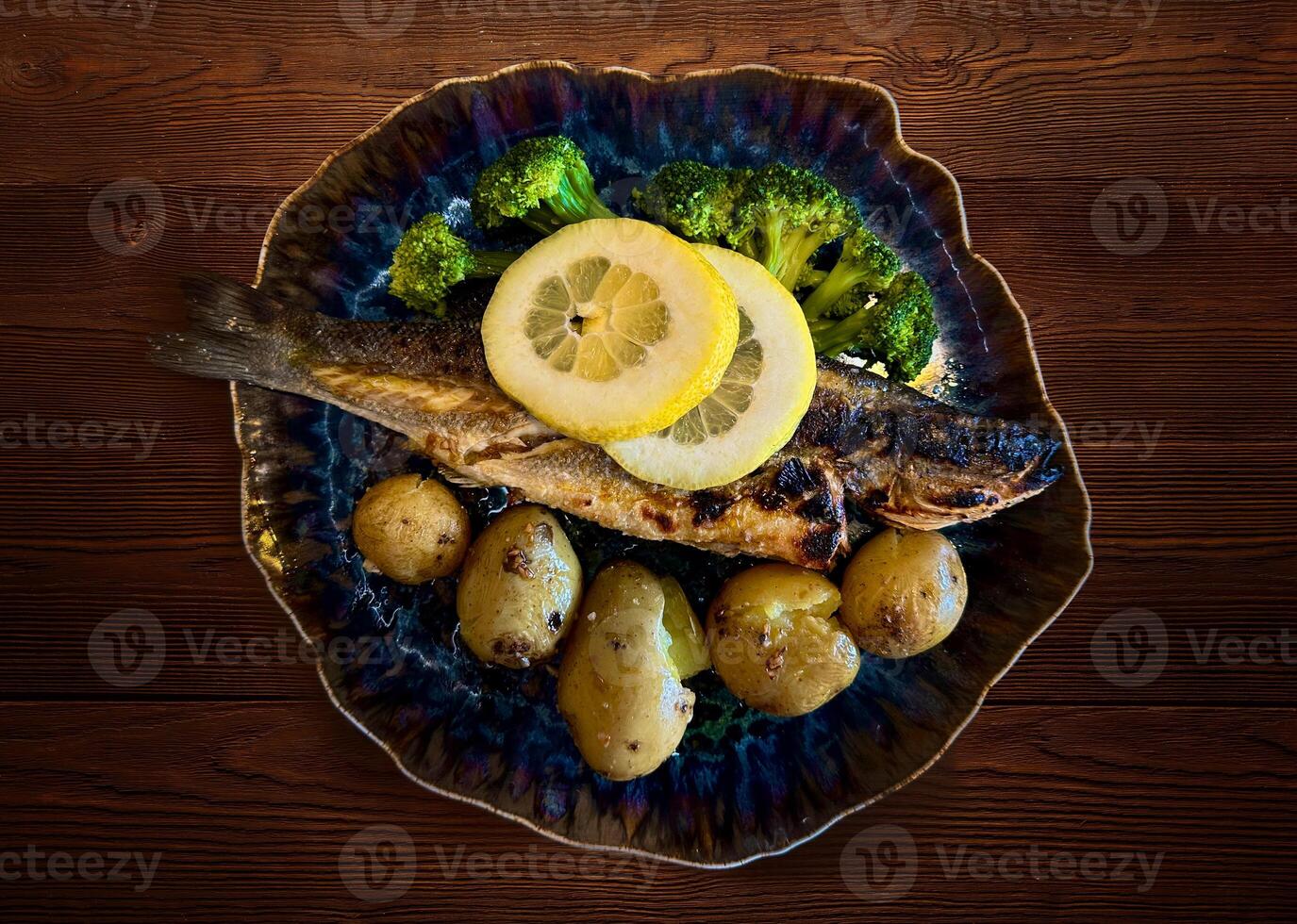 Golden roasted fish with potatoes, lemon and broccoli, served on wooden table. Overhead, horizontal - image photo