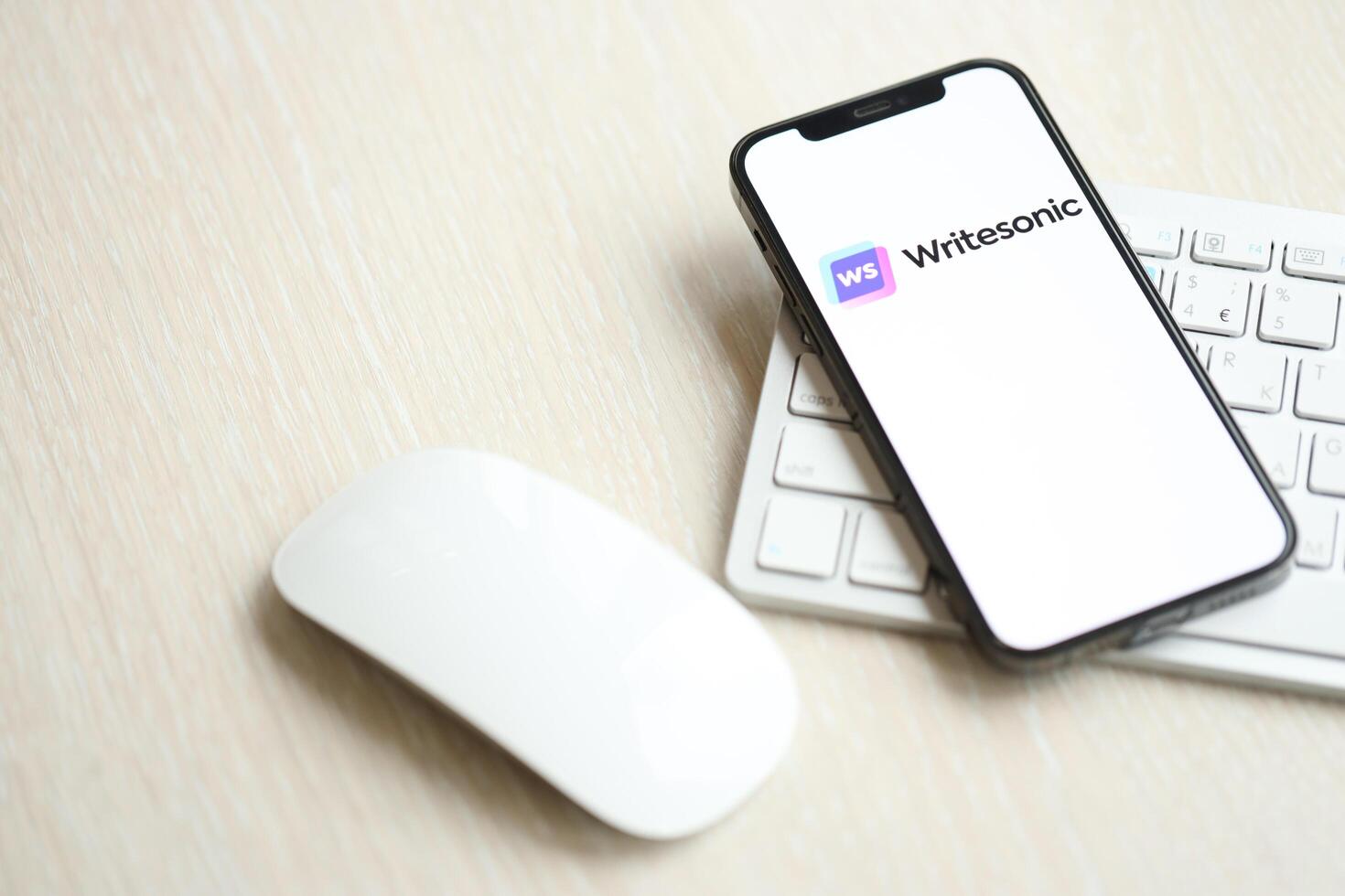 KYIV, UKRAINE - MARCH 17, 2024 Writesonic logo on iPhone display screen with apple keyboard and mouse on table photo