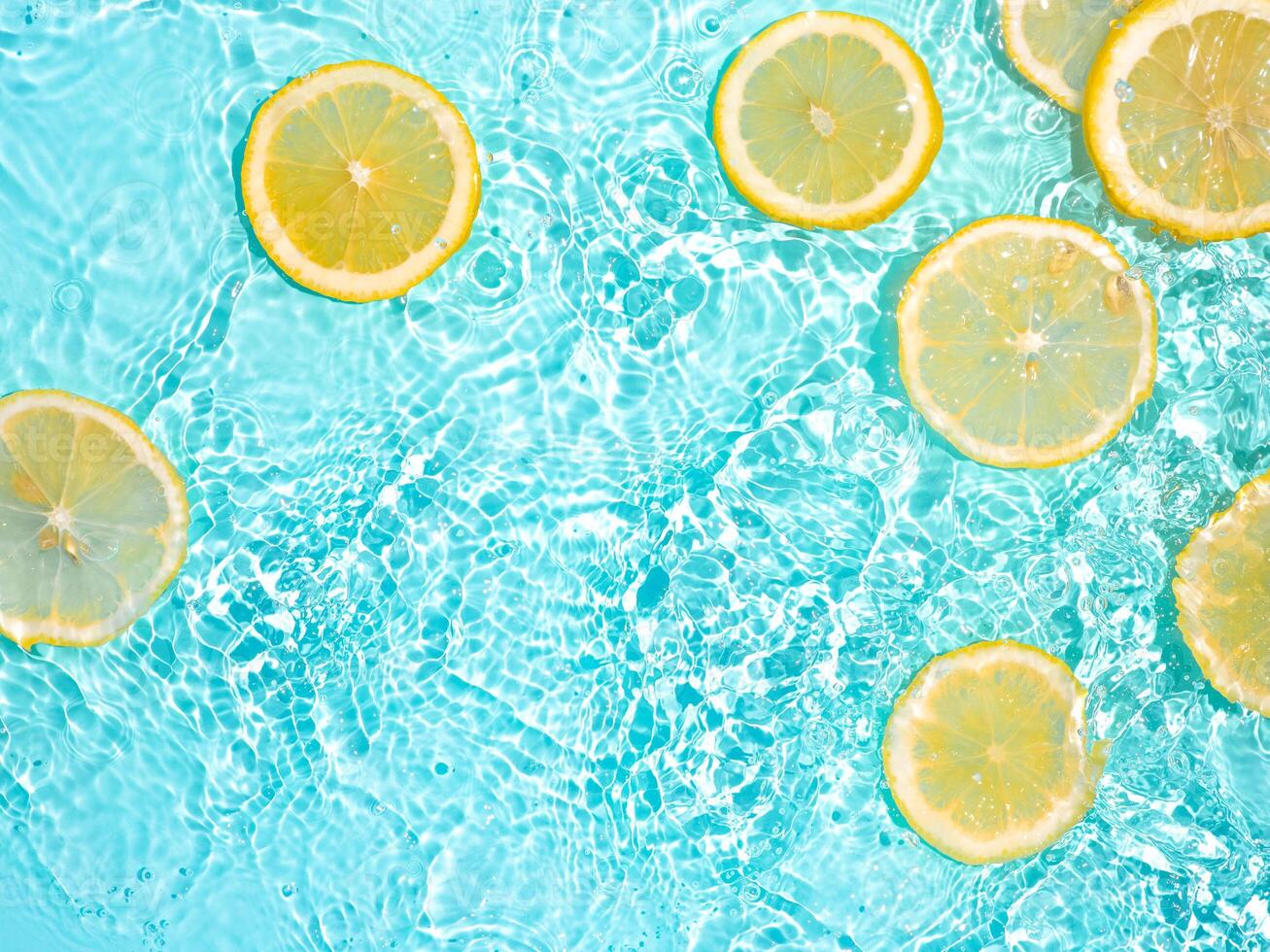 Lemon slices in clean transparent water over blue background with copy space. Water splashing on blue water surface in sunlight. Top view or flatlay. Summer, vacation, healthy eating concept photo