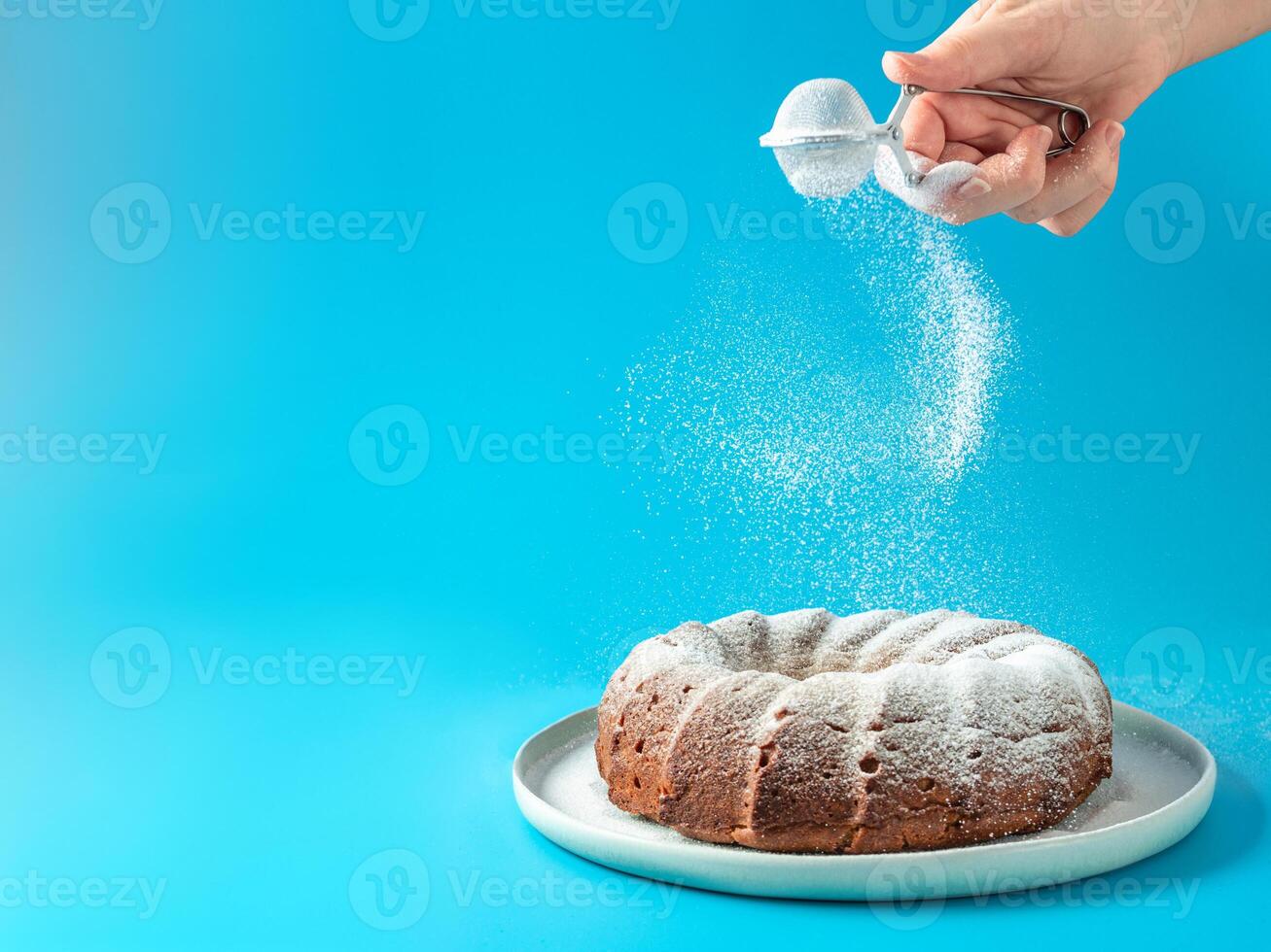 Woman's hand sprinkling icing sugar over fresh home made bundt cake. Powder sugar falls on fresh perfect bunt cake over blue background. Copy space for text. Ideas and recipes for breakfast or dessert photo