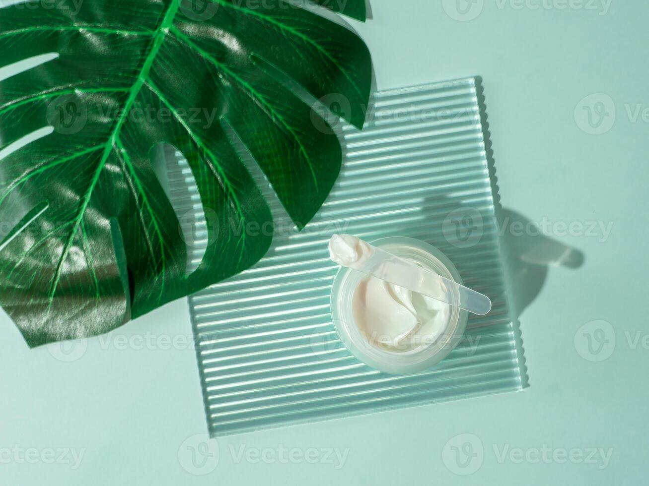 Cosmetic cream or moustirizer on transparent ribbed acrylic plate, tropical palm monstera leaf over blue background. Open round jar, aesthetic swirls cream and spatula. Top view flatlay. Copy space photo