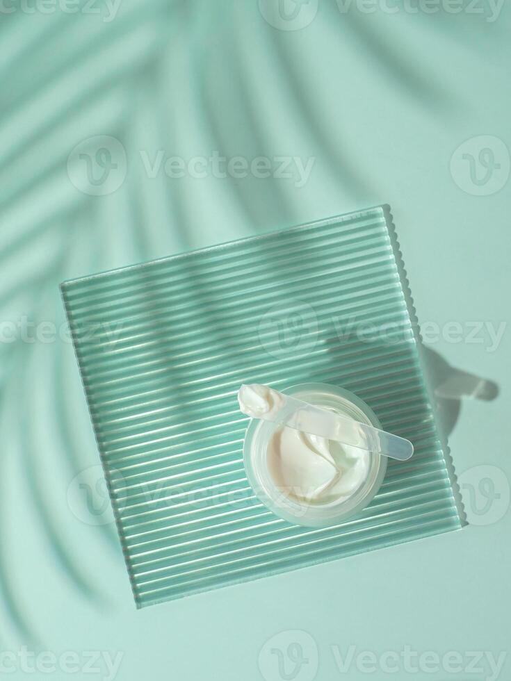 Cosmetic cream moustirizer, transparent ribbed acrylic plate, tropical palm leaf shadows on blue background. Open round glass jar,aesthetic swirls cream,spatula. Top view flatlay. Copy space. Vertical photo