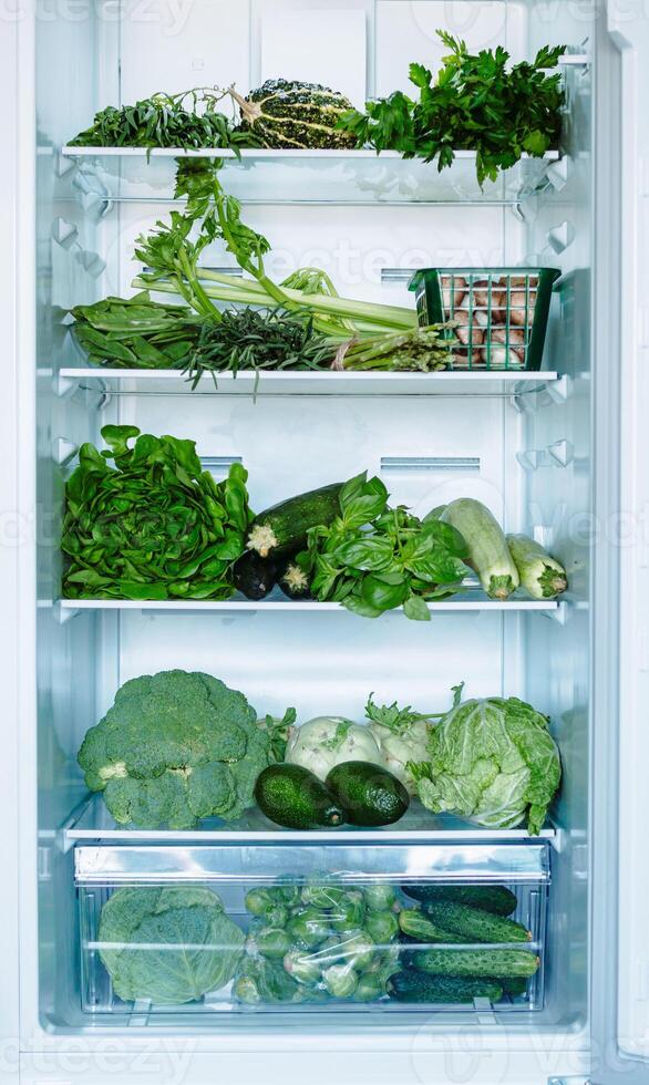 Fresh green vegetables in household refrigerator. Open full refrigerator with selected green vegetables and greens. Vertical shot of cabbages, zucchini, basil, avocado and cucumbers. Vegetarian food photo