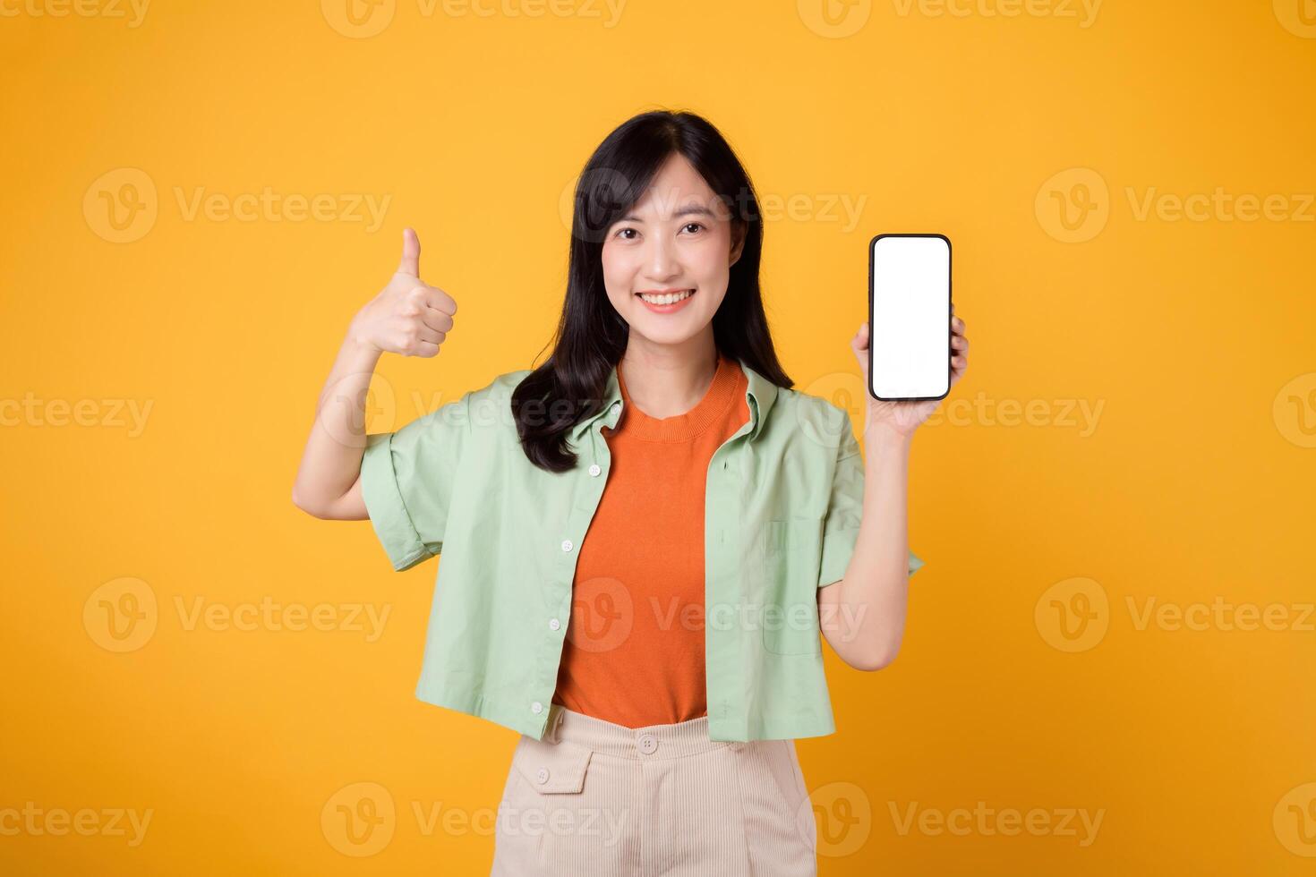 young Asian woman in her 30s, wearing orange shirt and green jumper, shows smartphone screen display with a thumbs-up gesture on yellow studio background. New mobile application concept. photo