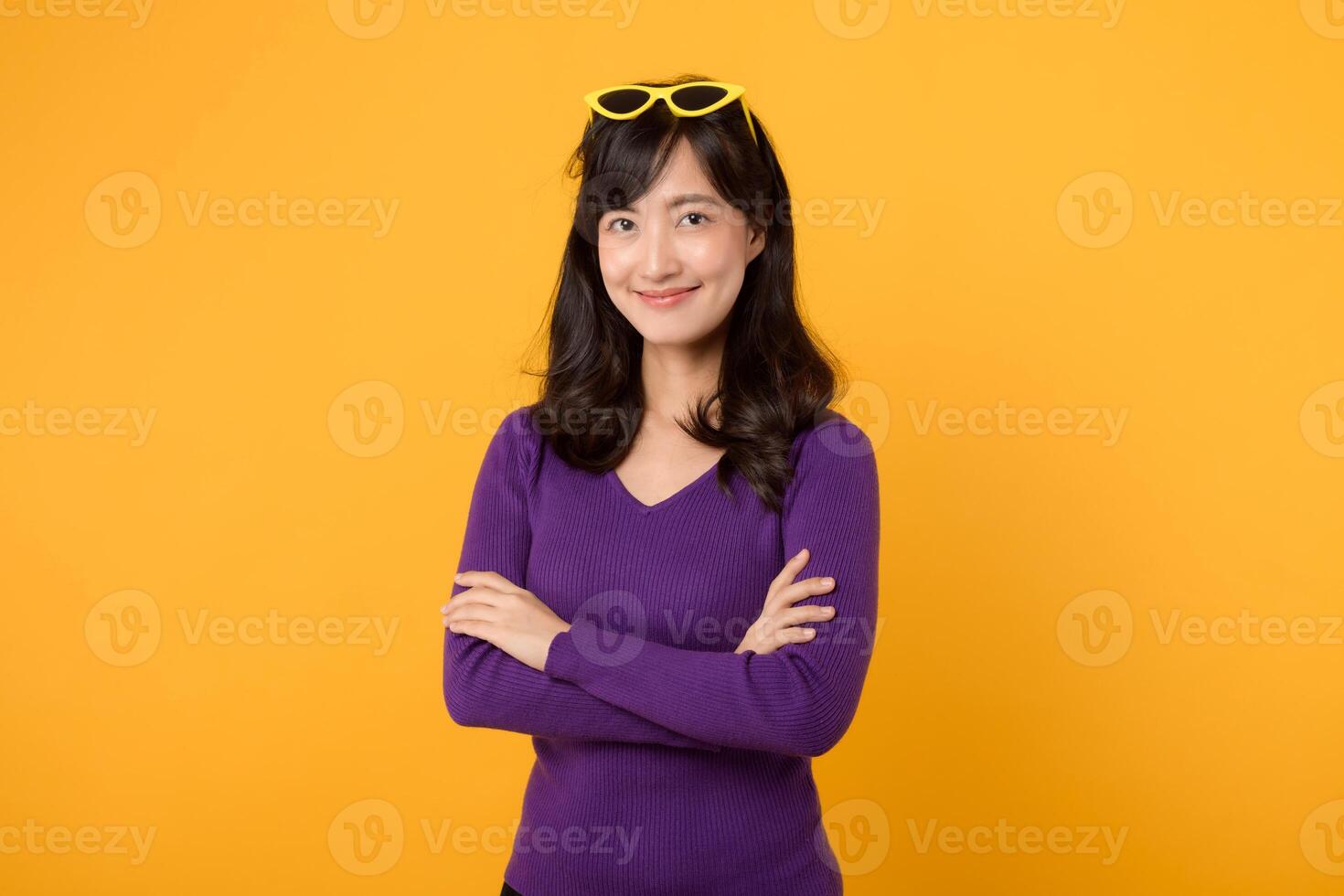 retrato de un confidente sonriente 30s asiático mujer vistiendo púrpura saltador camisa en pie con brazos doblada en cofre y mirando a el cámara aislado terminado amarillo antecedentes. foto