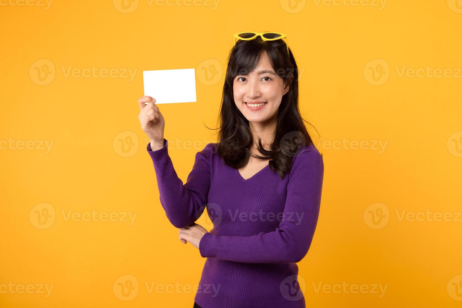 Young Asian woman 30s wearing purple sweater and yellow glasses showing and holding blank white card and holding her hip isolated on yellow background photo