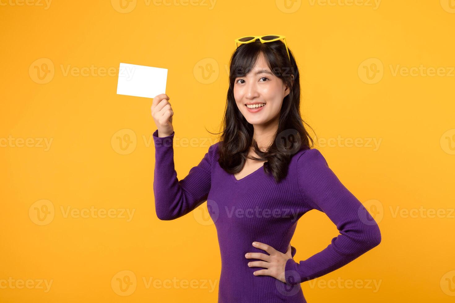 Young Asian woman 30s wearing purple sweater and yellow glasses showing and holding blank white card and holding her hip isolated on yellow background photo