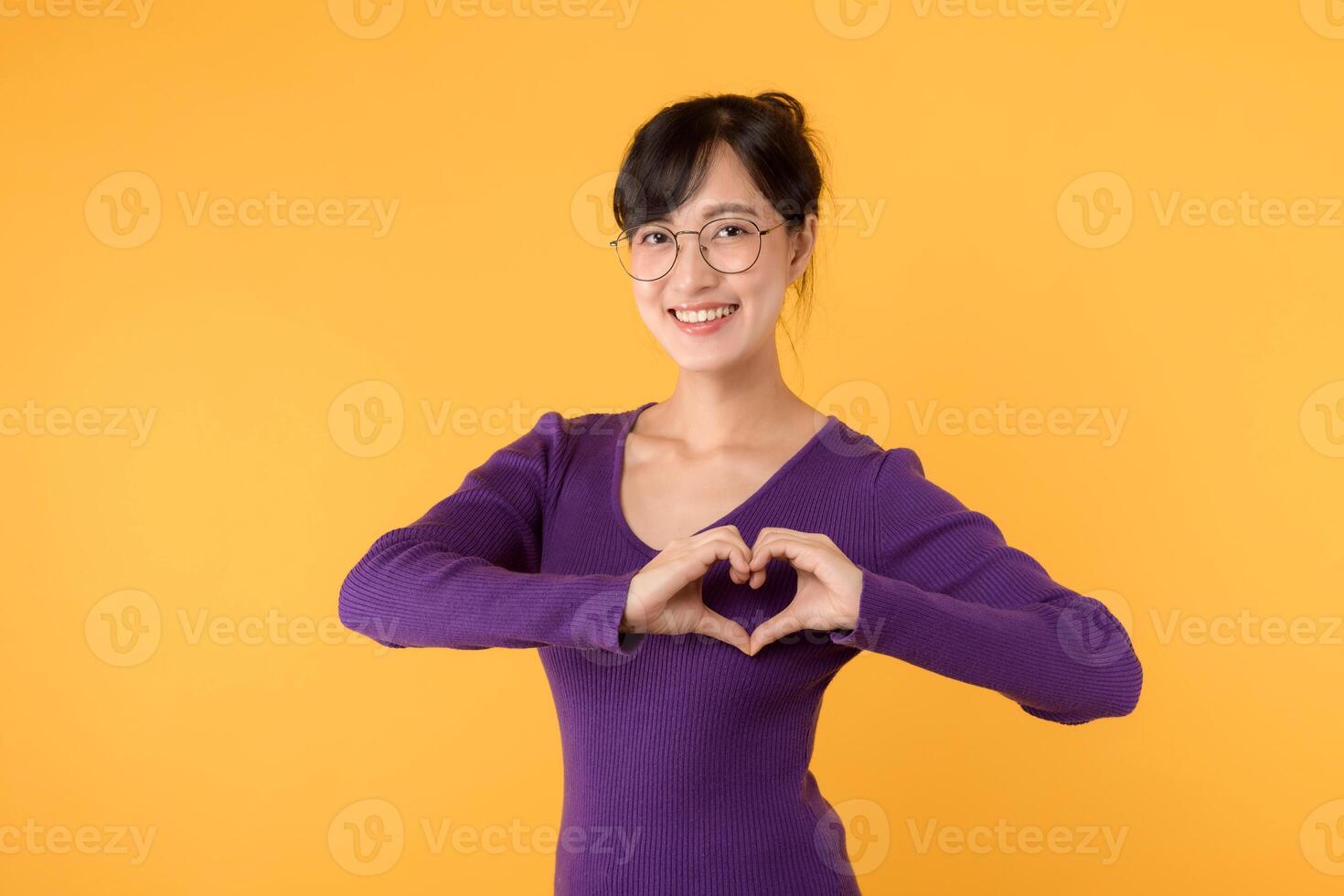 Portrait cheerful young Asian 30s woman happy smile wearing purple sweater shirt expressing heart hand sign gesture on chest isolated yellow background. Valentine romantic symbol advertising concept. photo
