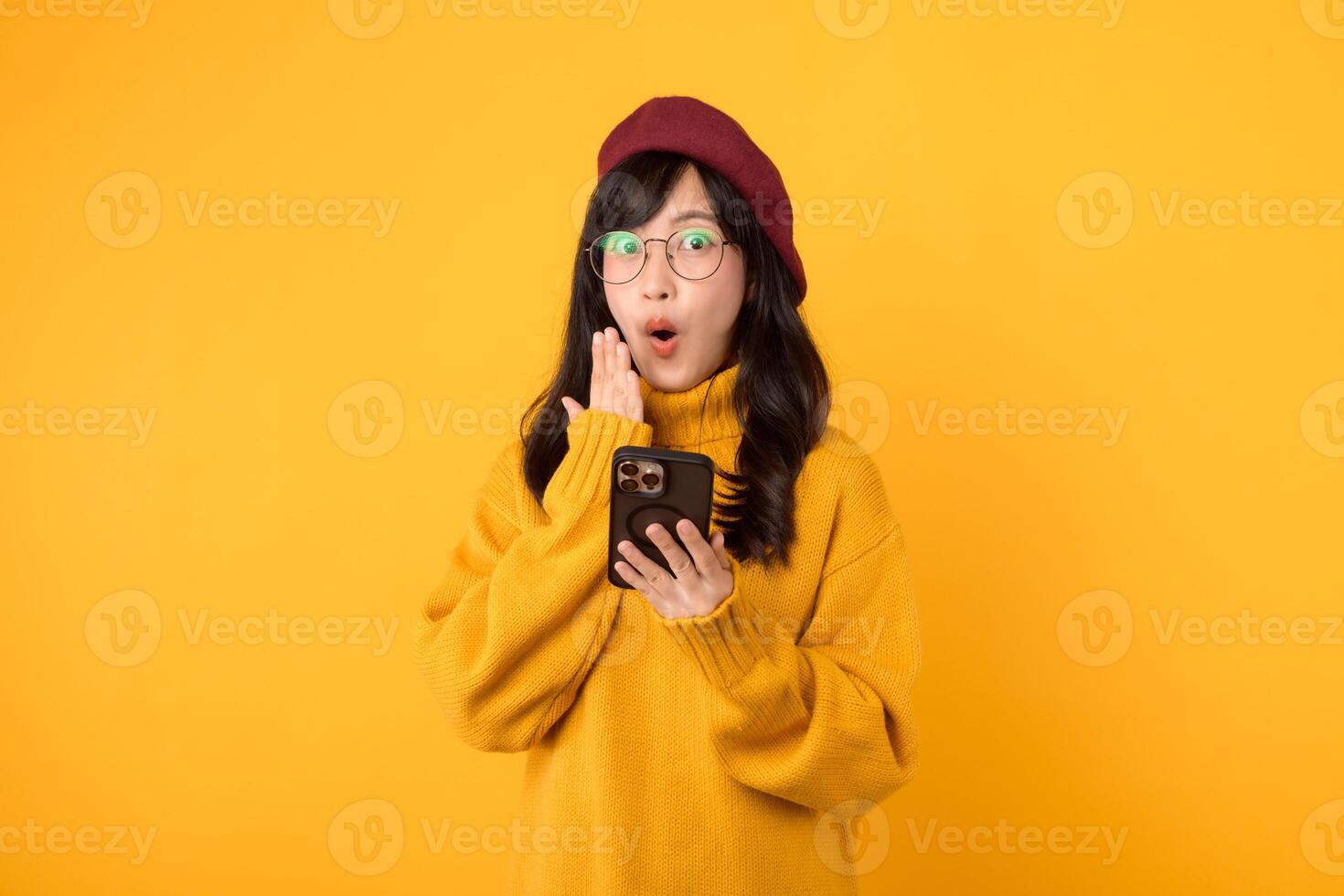 Surprised young asian 30s woman using mobile phone with positive expression, smiles broadly, dressed in yellow shirt and standing isolated on yellow background. Happy adorable glad rejoices success. photo