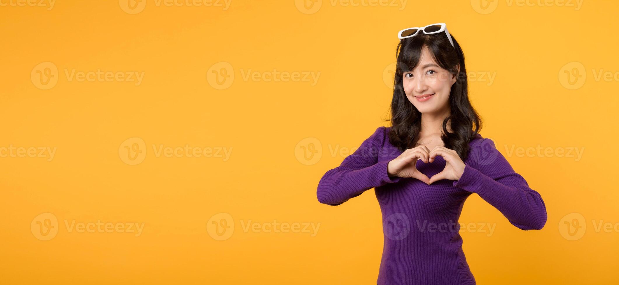 Young cheerful asian woman 30s wearing purple sweater dress and sunglasses shapes heart gesture on yellow background. photo
