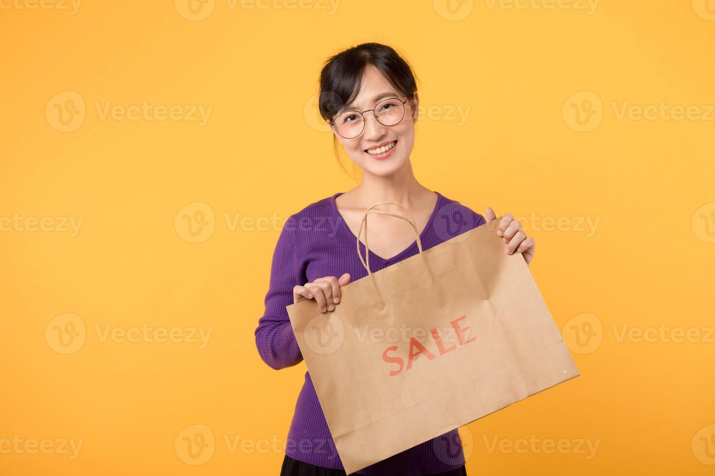 Asian young beautiful woman 30s wearing purple shirt holding paper shopping bag, presenting environmental sustainable packages, smiling with happiness isolated on yellow background with copy space. photo