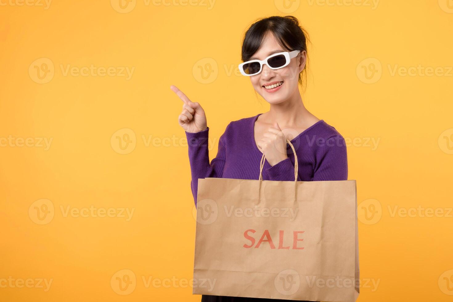 asiático joven mujer 30s vistiendo púrpura camisa y Gafas de sol participación papel compras bolsa, presentación ambiental sostenible paquetes, señalando dedo a gratis Copiar espacio. en contra amarillo antecedentes foto