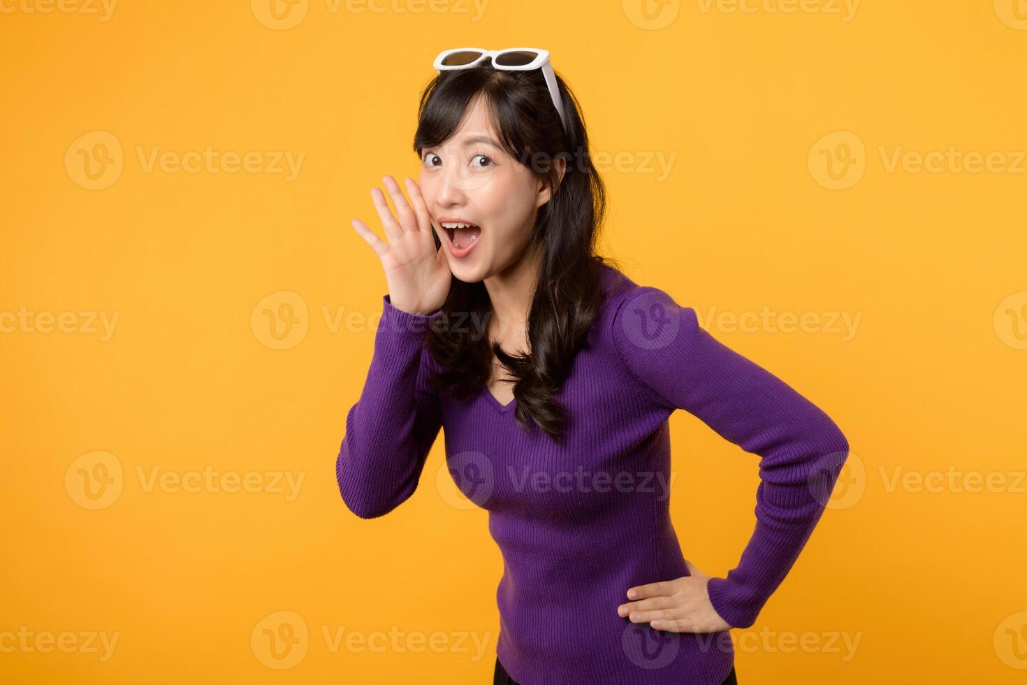 Portrait young beautiful asian woman 30s happy smile dressed in purple clothes shout to free space isolated on yellow studio background. Presenting product Beautiful girl act like telling to customer photo