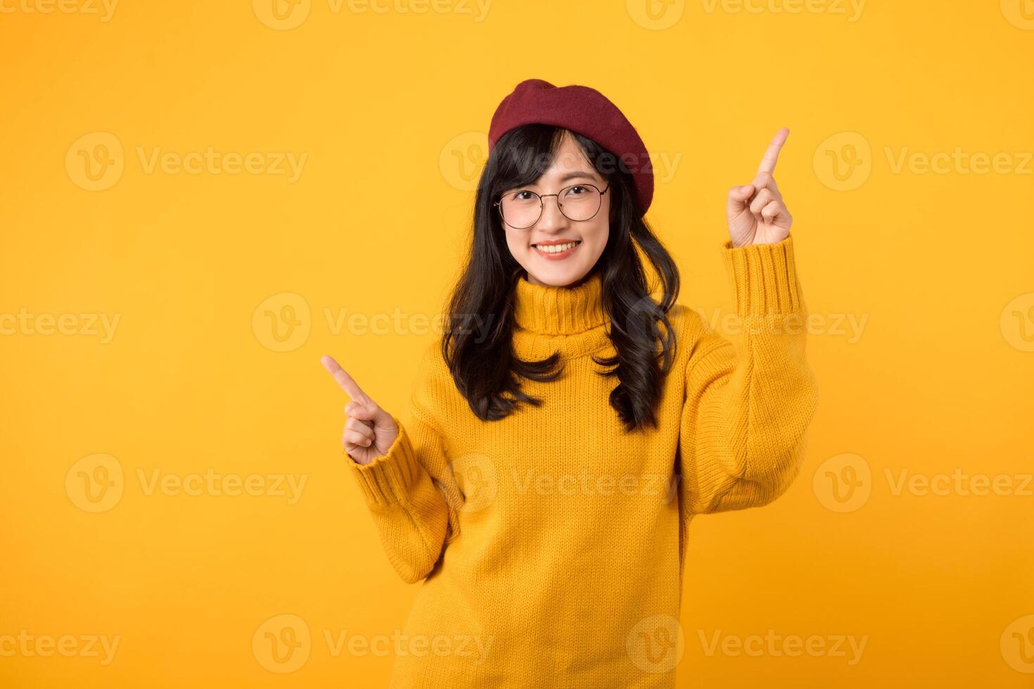 An elegant woman in her 30s, donning a red beret and eyeglasses, radiating happiness and pointing her finger to free copy space against a vibrant yellow background. photo