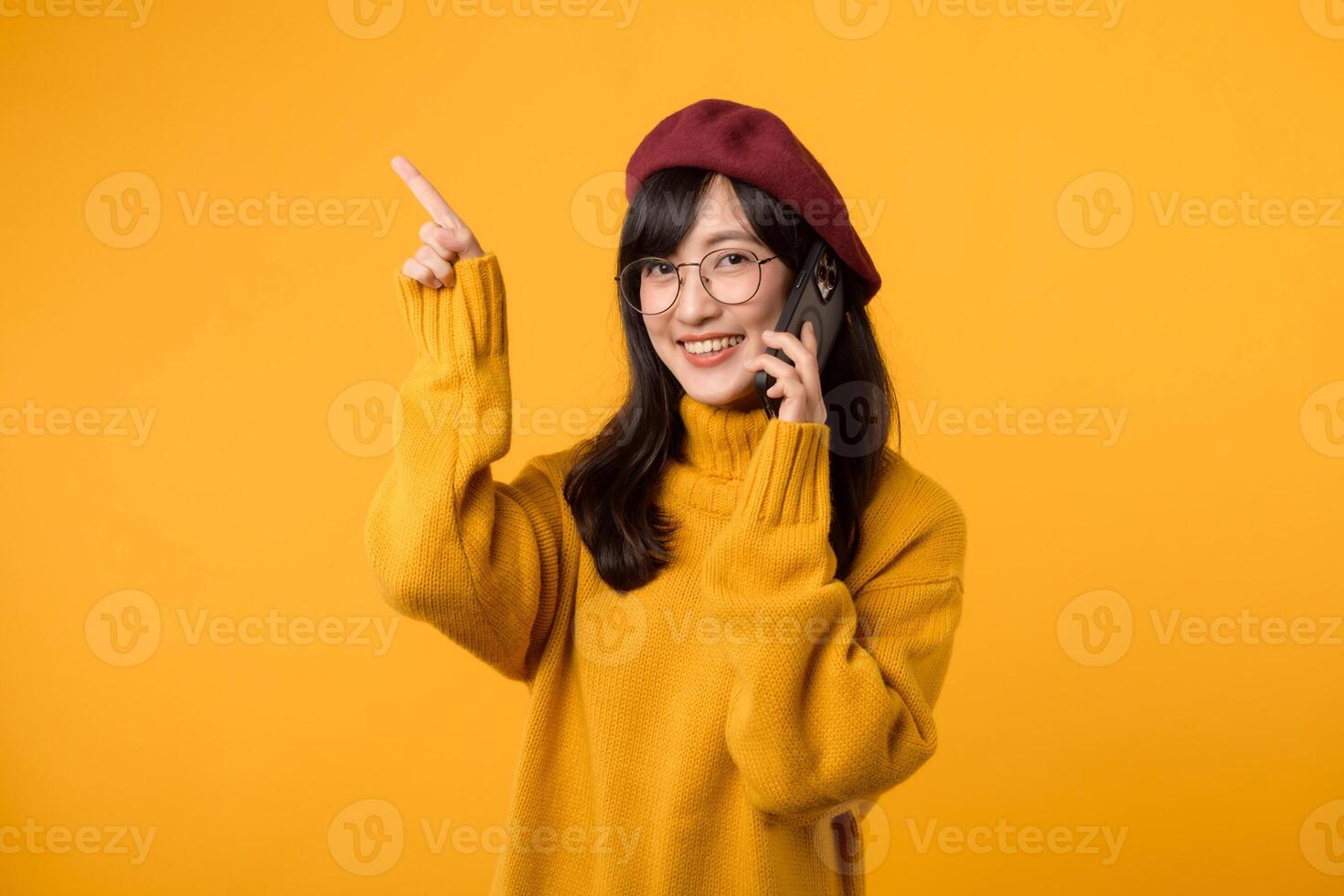 Female model in a vibrant beret and yellow sweater, pointing to free copy space on her mobile phone, set against a bright yellow background. photo