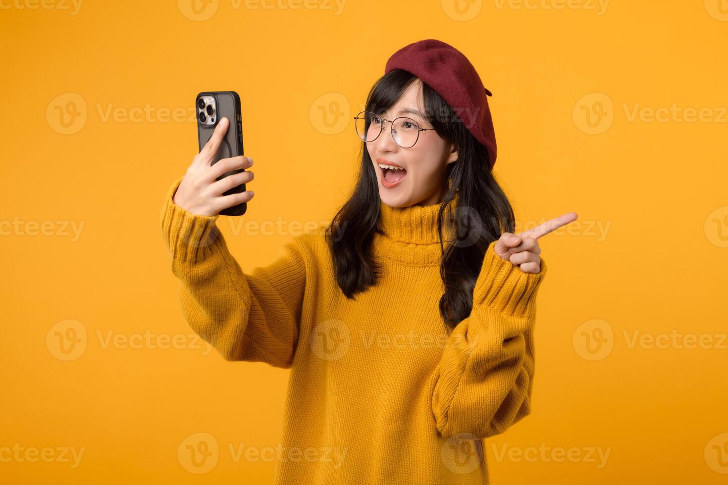 Portrait young beautiful 30s asian woman happy smile wearing yellow shirt and red hat pointing finger gesture to free copy space and using smartphone against yellow background. new application photo