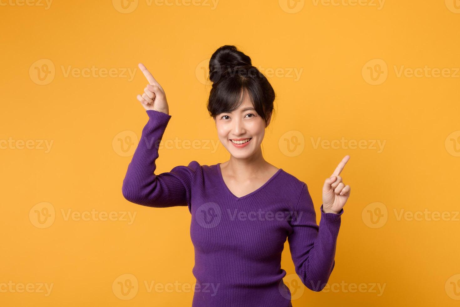 Beautiful young Asian woman 30s wearing purple shirt pointing up to copy space with smile face and happy. Pretty girl act like a satisfied product, use for advertising with yellow background photo