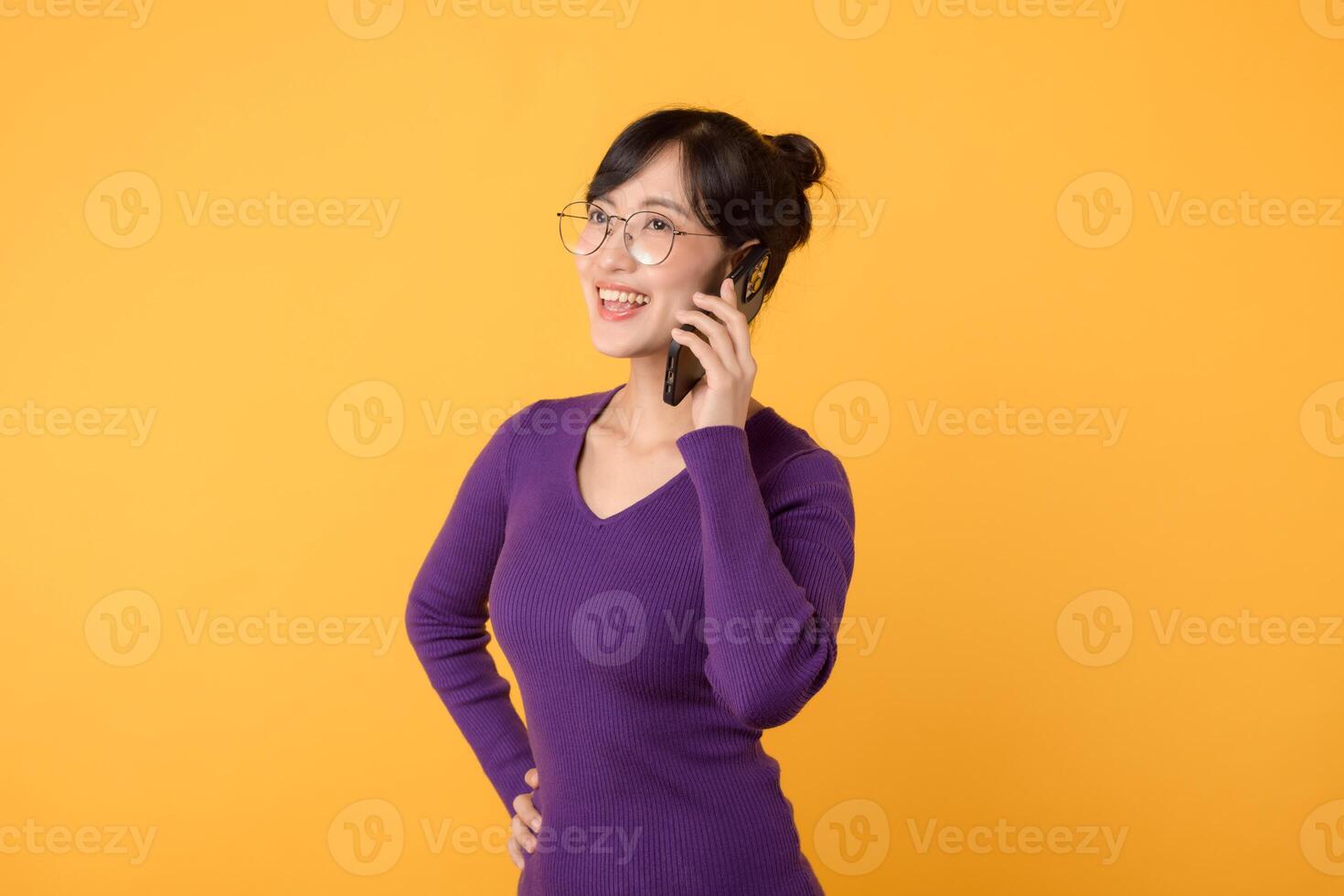 Vibrant Asian woman in her 30s, wearing purple shirt and sunglasses, using smartphone on vibrant yellow background. New mobile app concept. photo