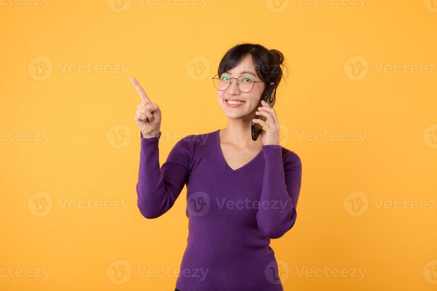 Experience the joy of online communication and shopping with a cheerful woman, wearing a purple shirt and eyeglasses, and her smartphone. Stay connected, stay smart against a yellow background photo