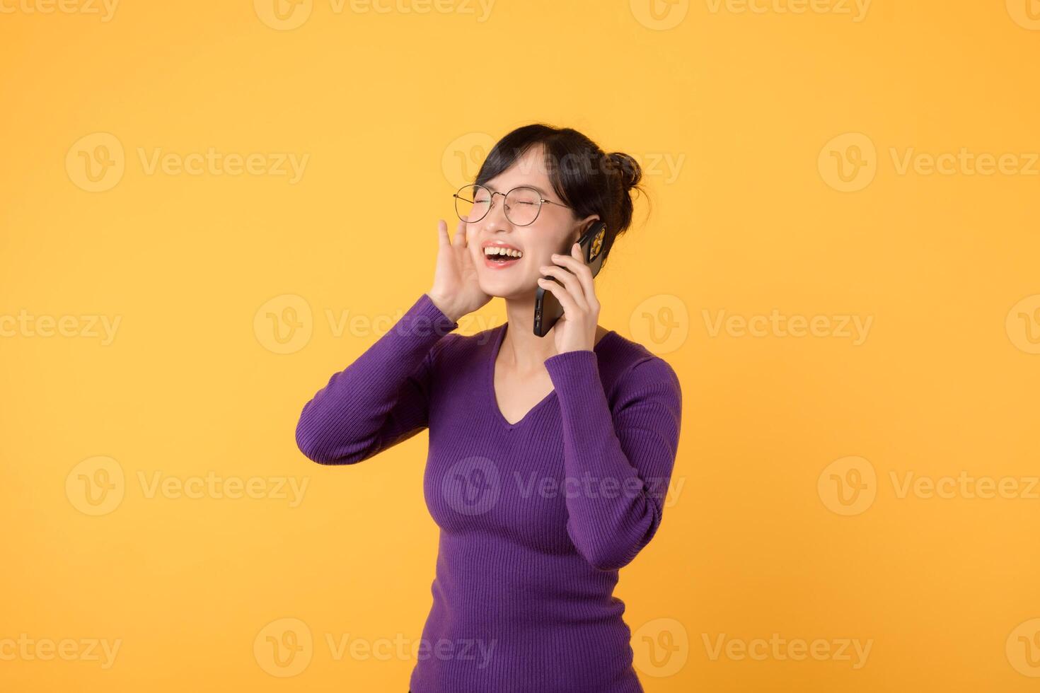 Portrait young Asian woman 30s wearing purple shirt and glasses receive an exited news from mobile phone isolated on yellow background. Great news concept. photo