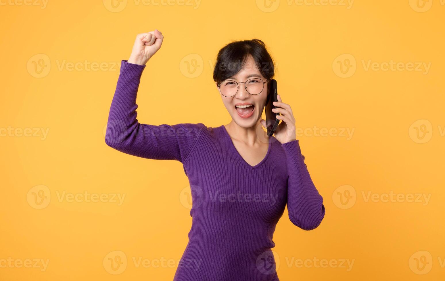 Portrait young Asian woman 30s wearing purple shirt and glasses receive an exited news from mobile phone isolated on yellow background. Great news concept. photo